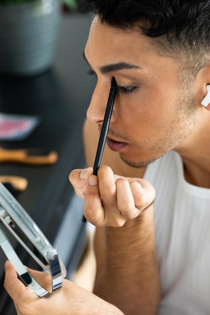 A man is carefully using a brow brush to fill in his eyebrows with a tinted brow gel. He is in a well-lit bathroom with a mirror in the background.