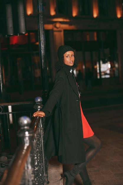 A person is attending an evening event wearing a black unisex oversized blazer layered over a sparkling gold sequin dress. They accessorize with high heels and a clutch. The setting is a softly lit urban scene.
