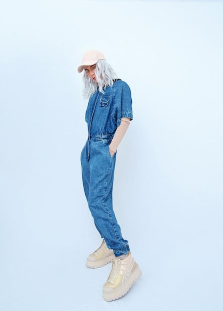 A person wearing a light-wash denim unisex jumpsuit, accessorized with a baseball cap, white sneakers, and a crossbody bag. They are walking in a park on a sunny day, holding a coffee cup. The overall look is relaxed and casual.
