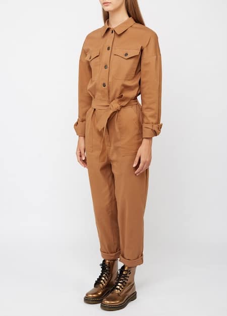 A person wearing a beige canvas unisex jumpsuit layered with a brown leather jacket and a colorful scarf. They are standing in an urban setting, showcasing the versatility of the jumpsuit for transitioning between seasons.