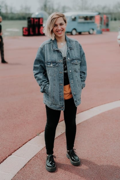 A person wearing an oversized, light-wash unisex denim jacket paired with slim-fit black jeans and white sneakers. The jacket has large pockets and a relaxed fit, emphasizing comfort and style. The person is standing against a minimalist, urban background.
