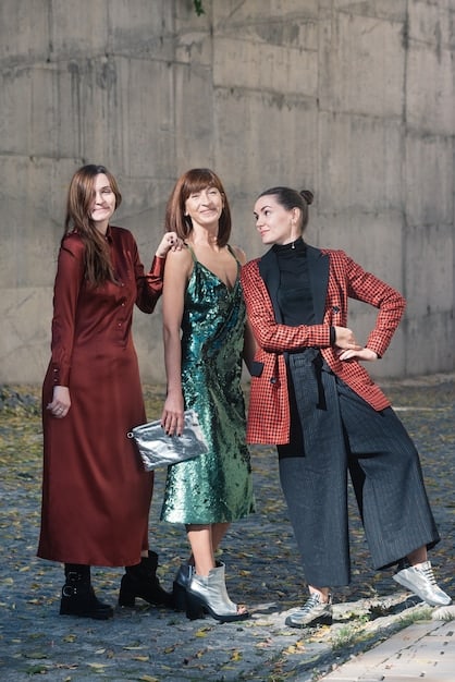 Three people standing side-by-side, each wearing an outfit featuring a different vibrant jewel tone. The first person wears an emerald green velvet blazer, the second person wears a sapphire blue silk shirt, and the third person wears amethyst purple wide-leg trousers. The background is a dark, moody setting.