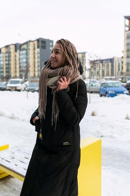 A person dressed in non-binary winter fashion, featuring a wool coat, corduroy pants, and a knitted scarf. The setting is an urban winter scene with a light snowfall.