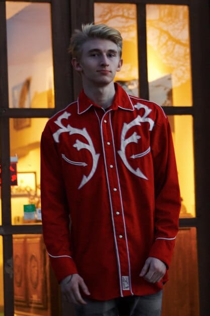 A person wearing a button-down shirt as a light outer layer over a graphic t-shirt, paired with tailored trousers and sneakers. The setting is a coffee shop, capturing a relaxed yet fashionable everyday street style.
