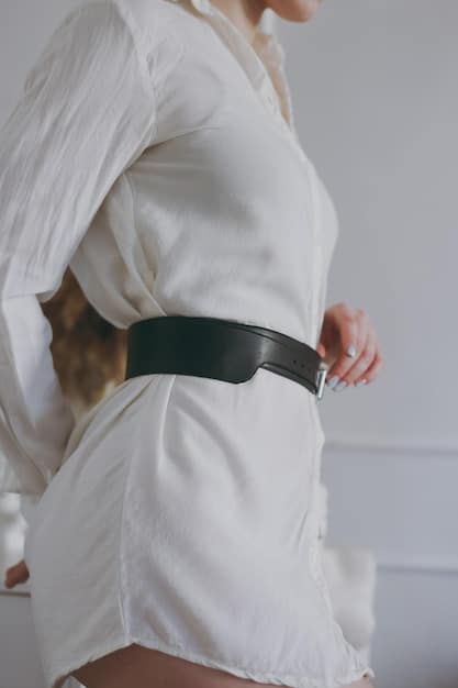 Close-up shot of a non-binary person adjusting a stylish belt worn over a jumpsuit, highlighting the detail and versatility of the accessory. The background is a blurred, modern interior space.