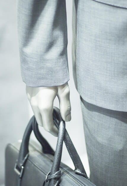 A structured bag in a neutral color (gray or beige) held by someone wearing a tailored suit. The bag features clean lines and minimal hardware, emphasizing its functional and stylish design.