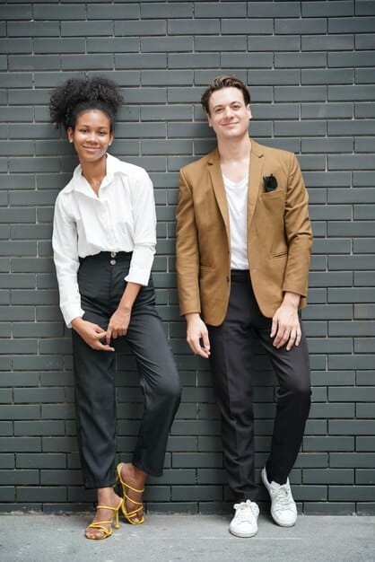Two people showcasing androgynous fashion: one wearing a perfectly tailored blazer and trousers, and the other in a loose but structured jumpsuit. They are posing in a studio with minimalist background, emphasizing the clothes' fit and style.