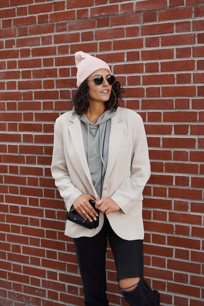 A person dressed in a light beige unisex blazer, a simple black crew-neck t-shirt, dark wash skinny jeans, and black Chelsea boots, standing against a brick wall.