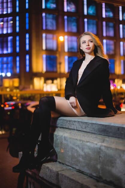 Close-up of a person wearing a black satin unisex blazer, a sequined top, leather pants, and high heels, with a blurred background of a dimly lit cocktail lounge.