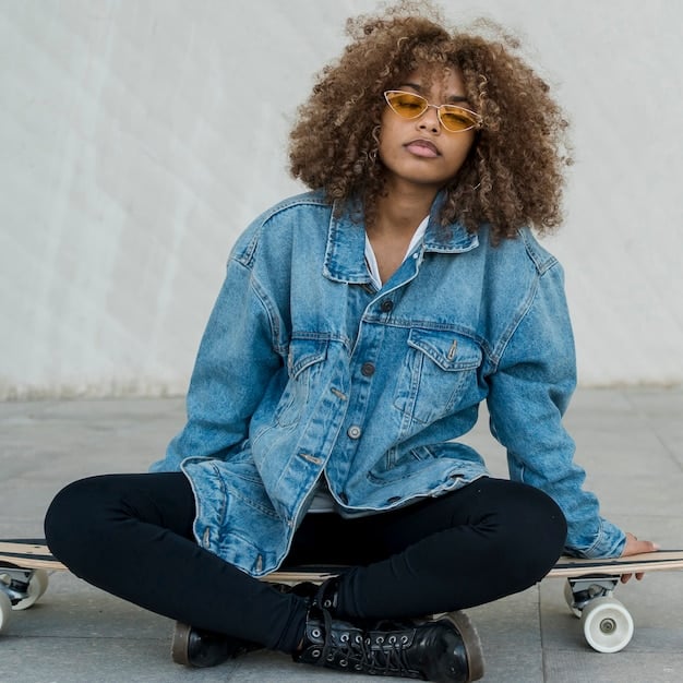A non-binary individual wearing a denim jacket over a hoodie, paired with black skinny jeans and combat boots. The individual is standing in front of a brick wall, with a confident and relaxed posture.