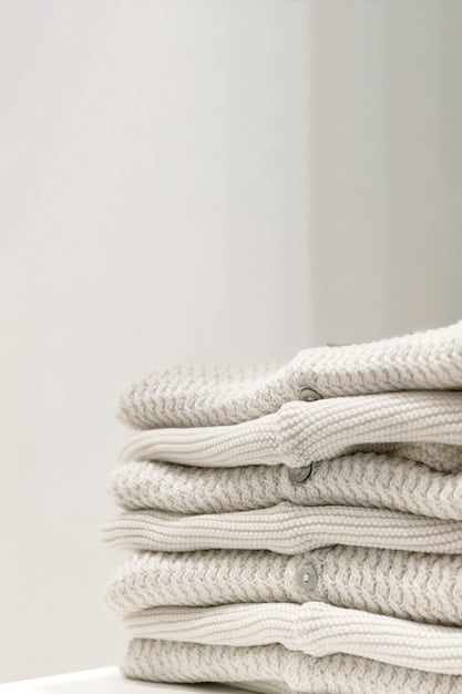 A close-up studio shot of a neatly folded pile of neutral-colored clothing, including a white button-down shirt, a grey sweater, black trousers, and a denim jacket. The lighting is soft and diffused, highlighting the texture and quality of the fabrics.