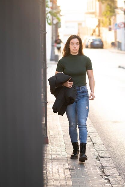 A person wearing a white t-shirt tucked into high-waisted jeans with a black belt. They are wearing black combat boots and a silver chain necklace. The background is an urban sidewalk.