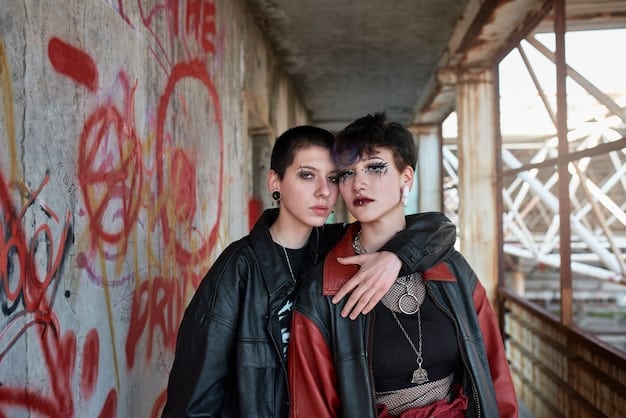 Two people are standing against a brick wall, both wearing androgynous layered outfits. One is wearing a neutral-toned ensemble with a pop of burgundy, while the other is wearing various shades of blue. The clothing items consist of blazers, sweaters, dress pants, and scarves.