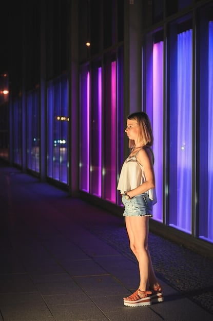 A full-length shot of a person standing confidently in a brightly lit urban setting. They are wearing a black turtleneck, wide-leg pinstripe trousers, and chunky combat boots. A silver chain hangs from their belt loop, and they have a short, styled haircut.