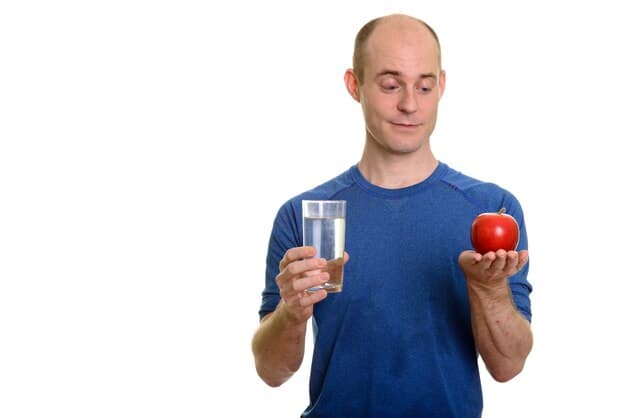 A close-up of a man holding a glass of water, with fresh fruits and vegetables in the background, emphasizing healthy hydration and diet.
