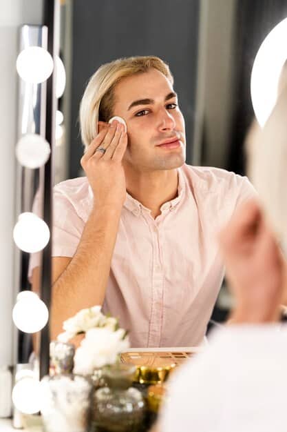 A man in a bathroom mirror carefully applying a small amount of concealer to a minor blemish on his cheek using his fingertip, focusing on precise, light application.