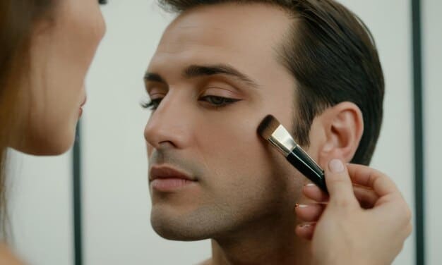 A man gently dabbing concealer under his eye with a small, tapered concealer brush, focusing on brightening the area. The lighting is soft, highlighting the natural application.