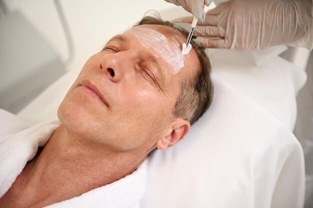 A close-up side profile of a man receiving a chemical peel from a dermatologist, with a gentle applicator applying the solution to his cheek. His expression is calm, indicating mild discomfort at most.