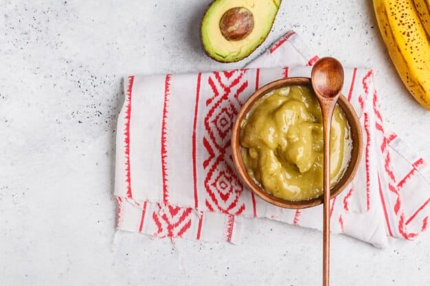 Overhead shot of a glass bowl filled with ripe avocado being mashed with a fork, alongside segments of banana and a drizzle of honey, suggesting a wholesome, natural skincare preparation.