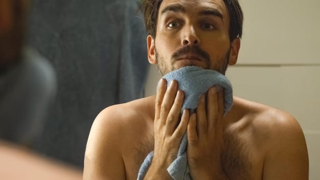 A close-up shot of a man's hand gently patting his face with a clean, soft white towel after cleansing. His skin looks fresh and dewy, and there are splashes of water nearby.