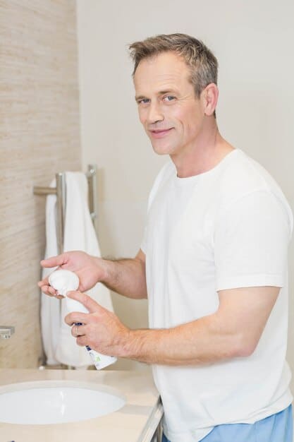 A man gently washing his face with a foaming cleanser, water splashing, set against a clean, bright bathroom background. The product bottle is generic but suggests a dermatologist-recommended type.