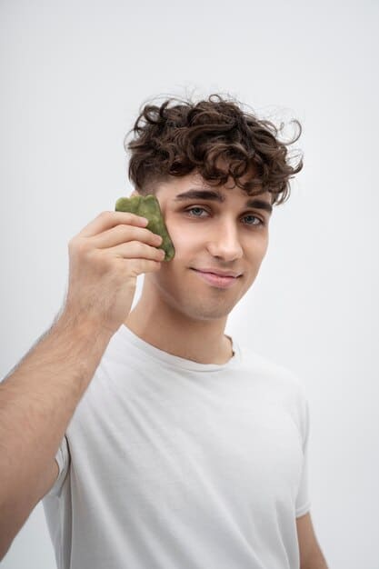 A man in his early 30s, with a healthy, natural complexion, subtly applying a green color-correcting concealer stick to a small red area on his cheek, demonstrating precise and discreet use. His expression is focused, and the background is a clean, well-lit bathroom vanity.
