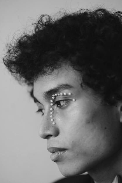 A close-up of a diverse male model with a striking, minimalist black graphic eyeliner design, featuring a sharp, angular line extending from the outer corner, set against a dark, moody background to emphasize the eye makeup.