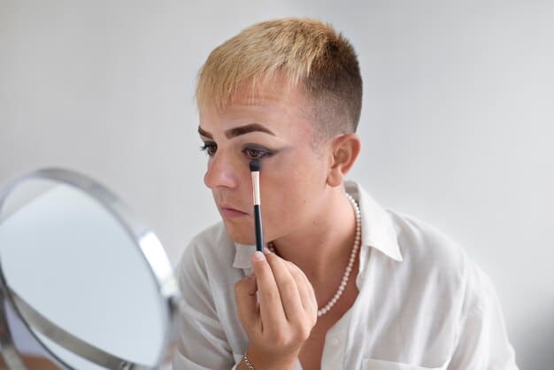 A man in his early 30s applying a liquid concealer with a doe-foot applicator under his eye, looking in a mirror. The background is a clean, modern bathroom with soft lighting, emphasizing a natural, everyday grooming ritual.