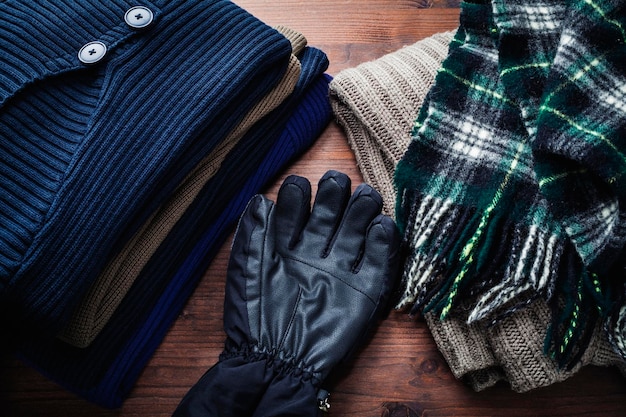 A close-up shot of various essential layering pieces for men. Items include a merino wool sweater, a quilted vest, a denim jacket, a flannel shirt, and a wool scarf, neatly arranged on a wooden surface. The color palette is dominated by earthy tones like olive green, navy, and charcoal gray. The Art of Layering: Mastering Men's Style for Fall and Winter.