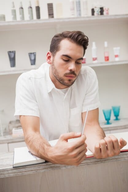 A man sitting in a professional nail salon, relaxed, as a manicurist gently files his nails. He is dressed in a stylish, casual outfit and appears comfortable and confident. This illustrates the acceptance and normalization of men receiving professional nail care as part of 'The Return of the Manicured Man: Nail Polish and Hand Care Trends for 2025'.