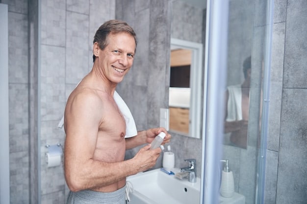 A man in his late 20s or early 30s is standing in his bathroom, smiling confidently as he applies cream in his bare face. He is looking directly at the camera.