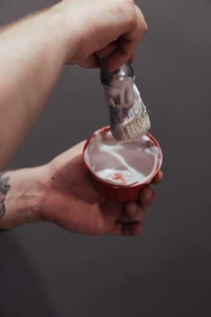 A hand holding a can or tube of Shaving Cream Y, with a shaving brush nearby. The background is a clean, modern bathroom setting. Water droplets suggest recent use.