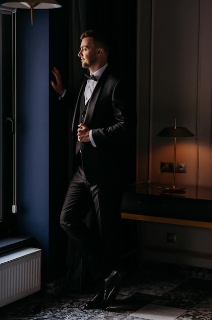 A sophisticated man dressed in a tailored suit stands in a modern, minimalist apartment, illuminated by natural light from a large window. He is spraying one of the new 2025 men's fragrances onto his wrist, his expression thoughtful as he evaluates the scent.  The background features clean lines, neutral colors, and subtle artistic details. A caption reads 