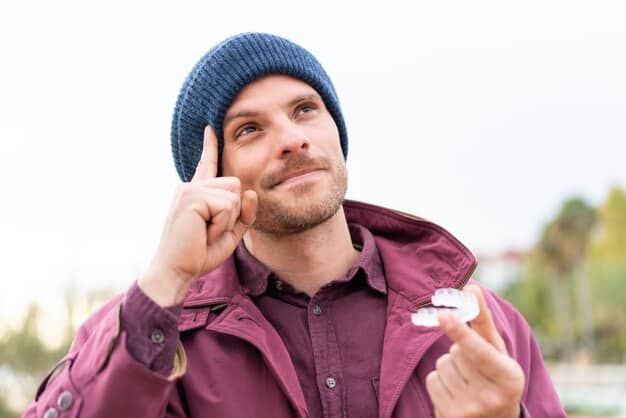 A man applying lip balm in a windy, outdoor setting. He's wearing casual clothing, and his expression suggests relief and comfort. The background shows a blurred landscape with trees and blowing leaves, emphasizing the need for lip protection in harsh conditions.