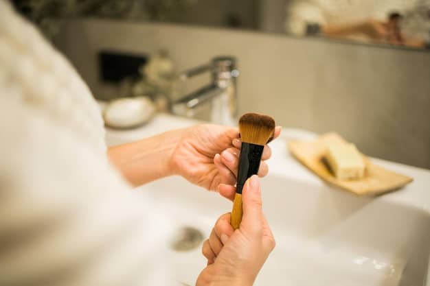 A close-up of a man's hand gently washing a clean makeup brush under a stream of lukewarm water, with a specialized brush cleaning mat visible. The scene emphasizes hygiene and careful maintenance.